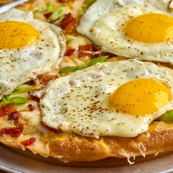 Close-up of a Mexican breakfast pizza topped with three fried eggs, bacon, cheese, and green onions.