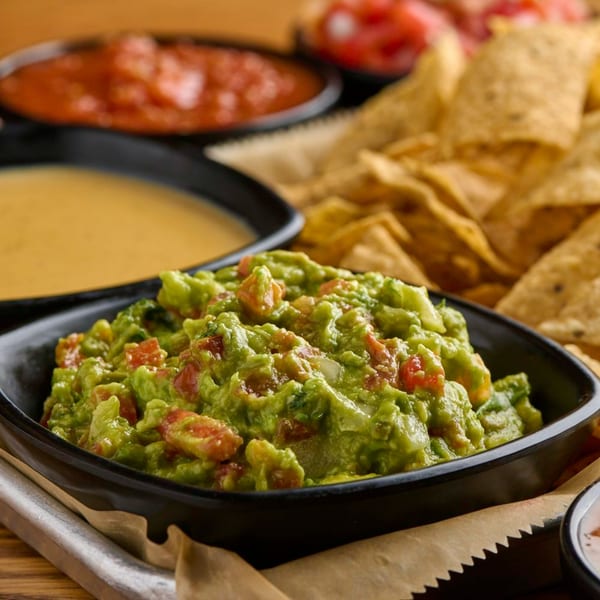 A black bowl of Salsaria's chunky guacamole with diced tomatoes, alongside tortilla chips, salsa, and queso.